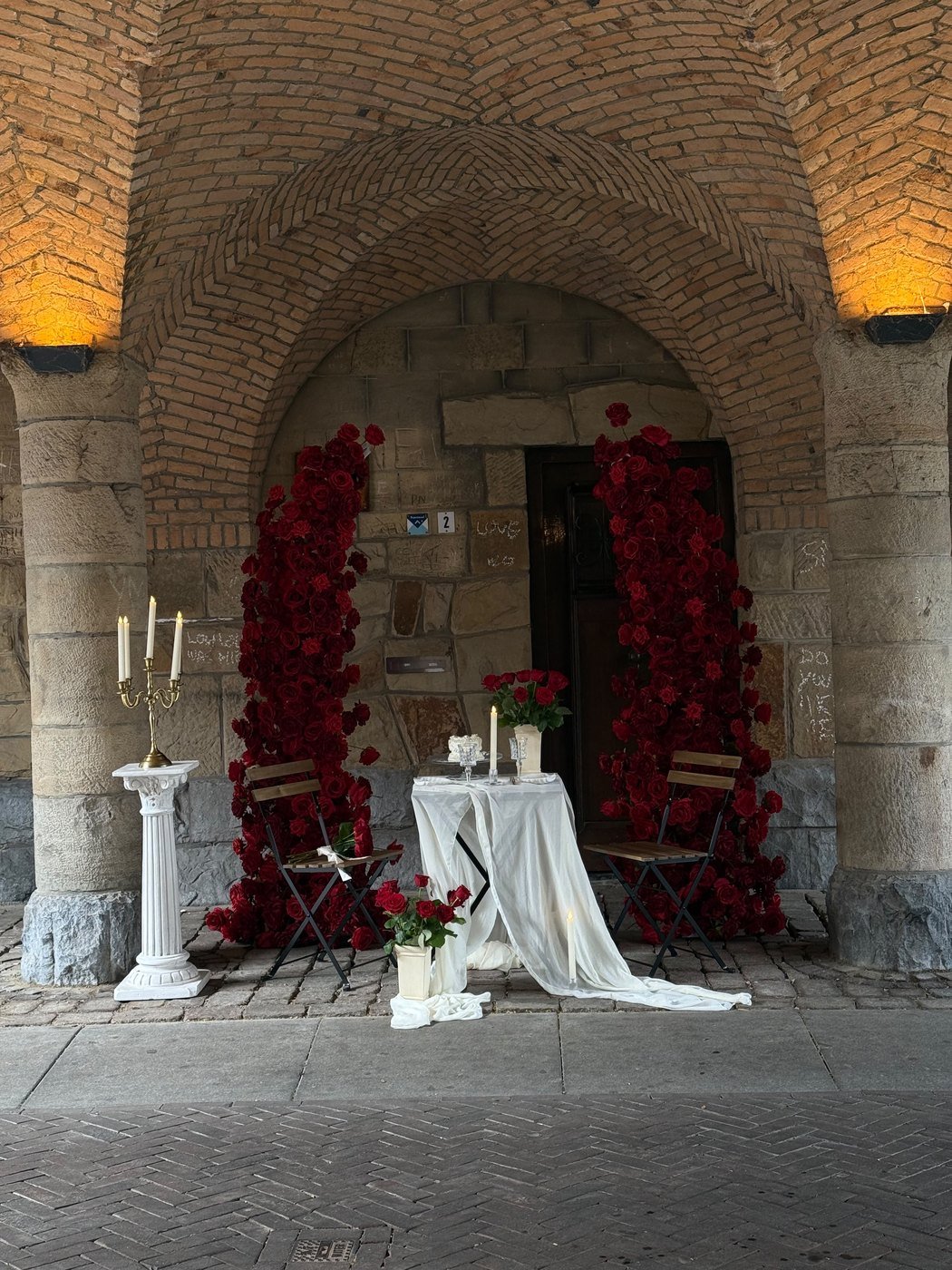 Huwelijksaanzoek onder een stenen poort, omlijst met een cascade van donkerrode rozen, met kandelaars en een klein gedekt tafeltje met twee stoelen ervoor