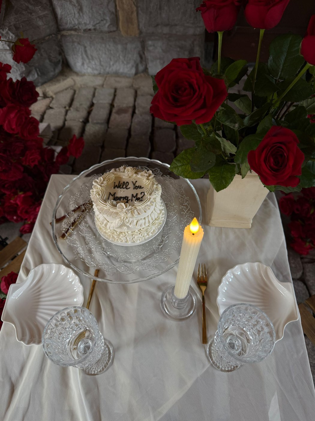 Detail van een huwelijksaanzoek bij kaarslicht, met een witte taart, donkerrode rozen, schelpvormige schaaltjes en kristallen glazen op wit linnen