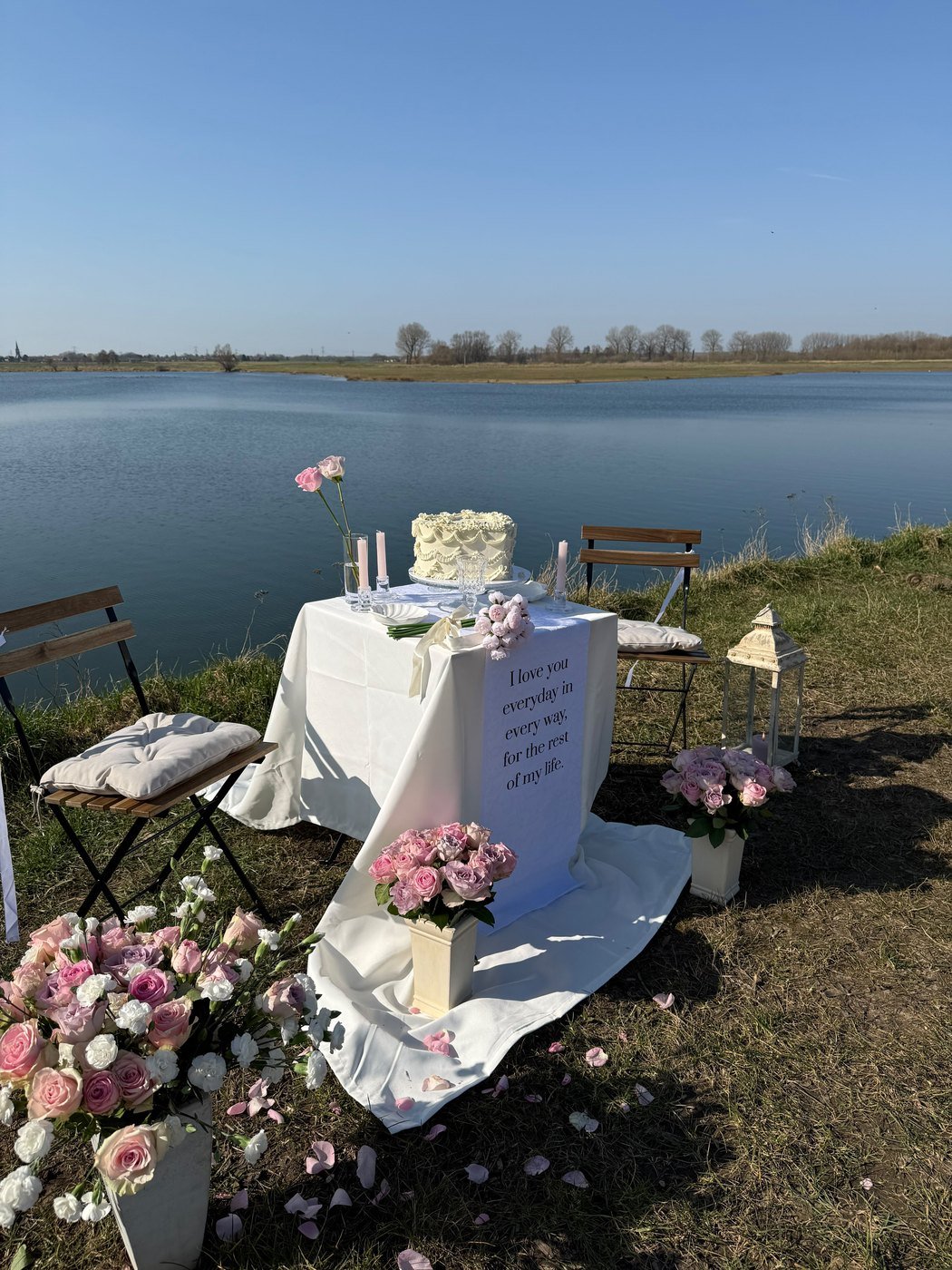 Huwelijksaanzoek aan de waterkant, met een wit gedrapeerde tafel, een taart, een bord met liefdesboodschap en volle bossen roze rozen op de grond